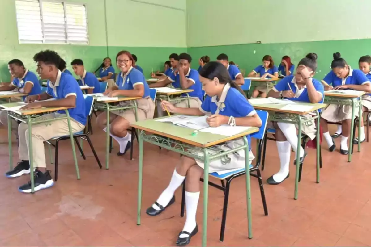 Estudiantes con uniforme escolar azul y beige sentados en pupitres dentro de un aula mientras realizan una actividad escrita Estudiantes con uniforme escolar azul y beige sentados en pupitres dentro de un aula mientras realizan una actividad escrita
