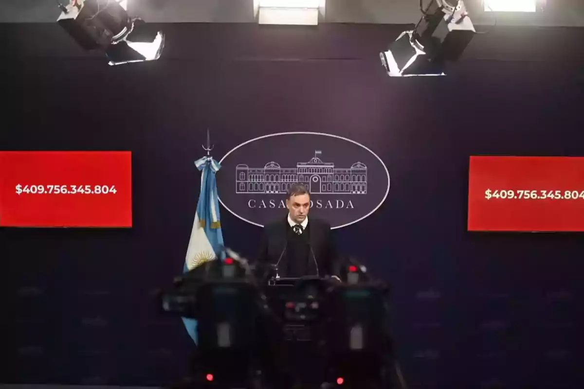 Un hombre de traje habla en una conferencia de prensa en la Casa Rosada de Argentina con dos pantallas rojas a los lados mostrando una cifra millonaria y cámaras grabando en primer plano. Un hombre de traje habla en una conferencia de prensa en la Casa Rosada de Argentina con dos pantallas rojas a los lados mostrando una cifra millonaria y cámaras grabando en primer plano.