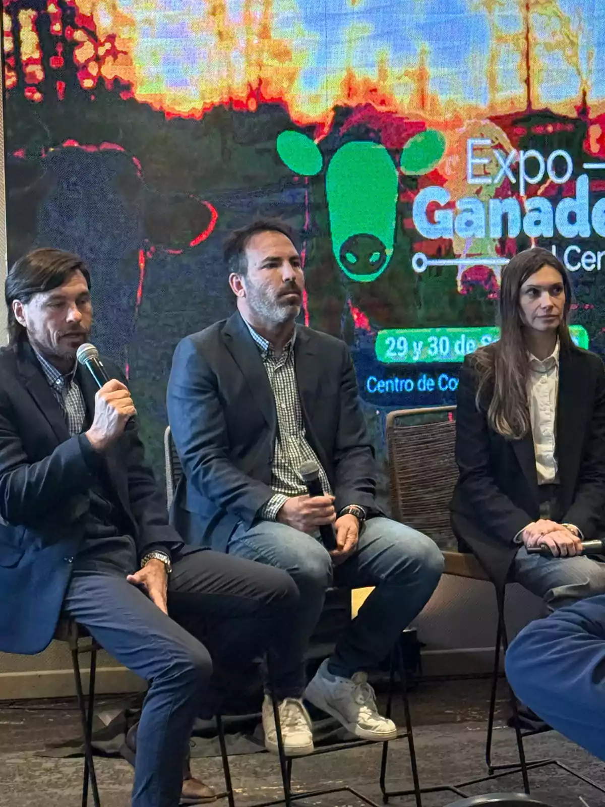 Tres personas sentadas en sillas durante un panel en un evento ganadero, dos de ellas sostienen micrófonos y al fondo se observa una pantalla con el logo de Expo Ganadera Tres personas sentadas en sillas durante un panel en un evento ganadero, dos de ellas sostienen micrófonos y al fondo se observa una pantalla con el logo de Expo Ganadera