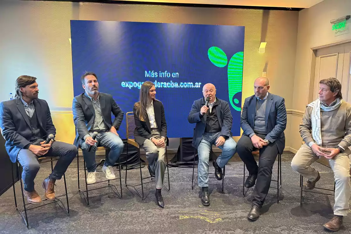 Seis personas sentadas en sillas durante un panel o conferencia, todas vestidas de manera formal, con un fondo de pantalla azul que muestra información de un sitio web. Seis personas sentadas en sillas durante un panel o conferencia, todas vestidas de manera formal, con un fondo de pantalla azul que muestra información de un sitio web.