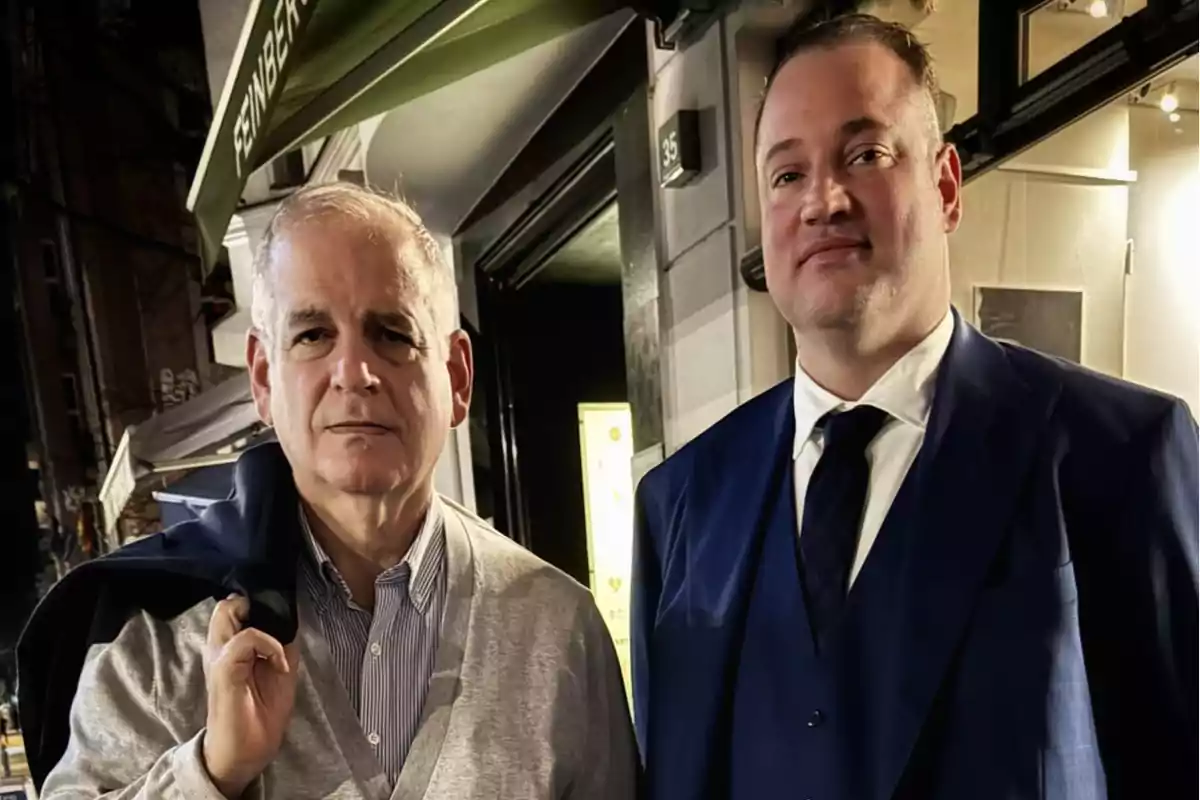 Dos hombres posan juntos en la entrada de un restaurante por la noche uno lleva un suéter gris y el otro viste un traje azul Dos hombres posan juntos en la entrada de un restaurante por la noche uno lleva un suéter gris y el otro viste un traje azul