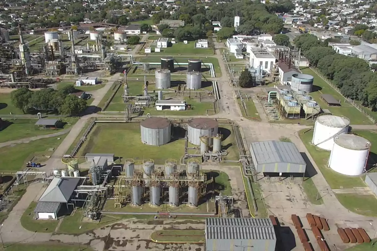 Aerial view of an industrial plant with several storage tanks, metal structures, and buildings surrounded by green areas and internal roads.