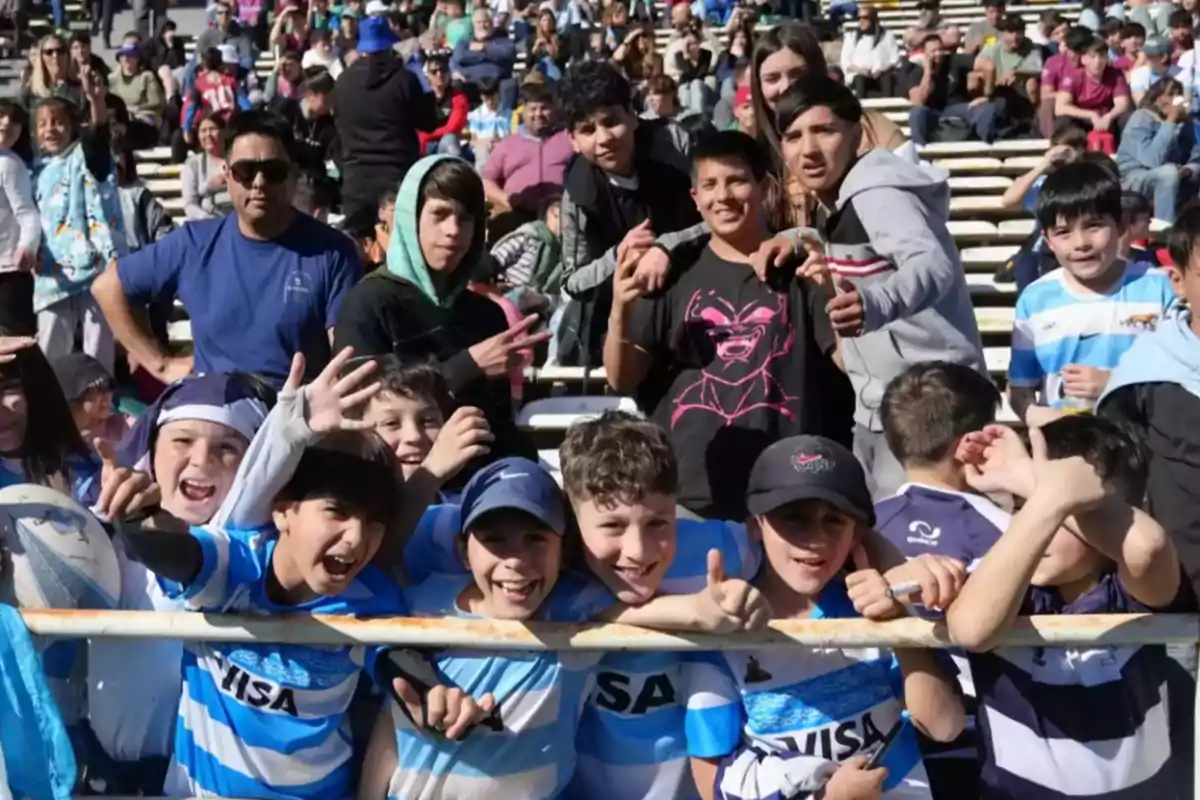 A group of children and teenagers smile and pose for the camera in a crowded sports stand, some wearing light blue and white rugby jerseys.