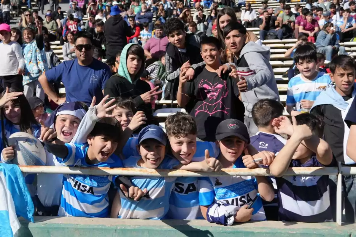 Un grupo de niños y adolescentes sonríen y posan para la cámara en las gradas de un estadio durante un evento deportivo, algunos llevan camisetas de rugby y hay público en el fondo.