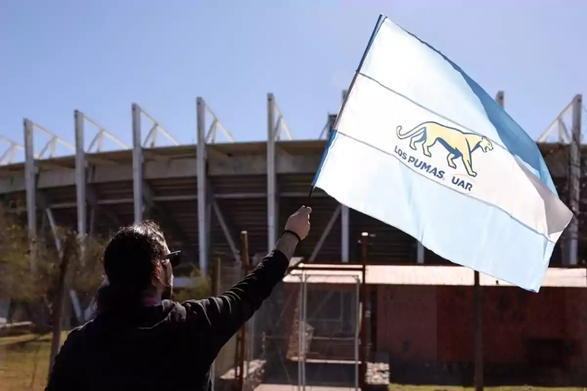 Persona de espaldas ondeando una bandera de Los Pumas UAR frente a un estadio