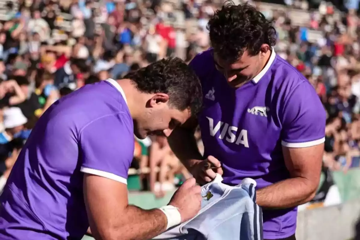 Two rugby players in purple jerseys sign a light blue jersey in a packed stadium. Two rugby players in purple jerseys sign a light blue jersey in a packed stadium.