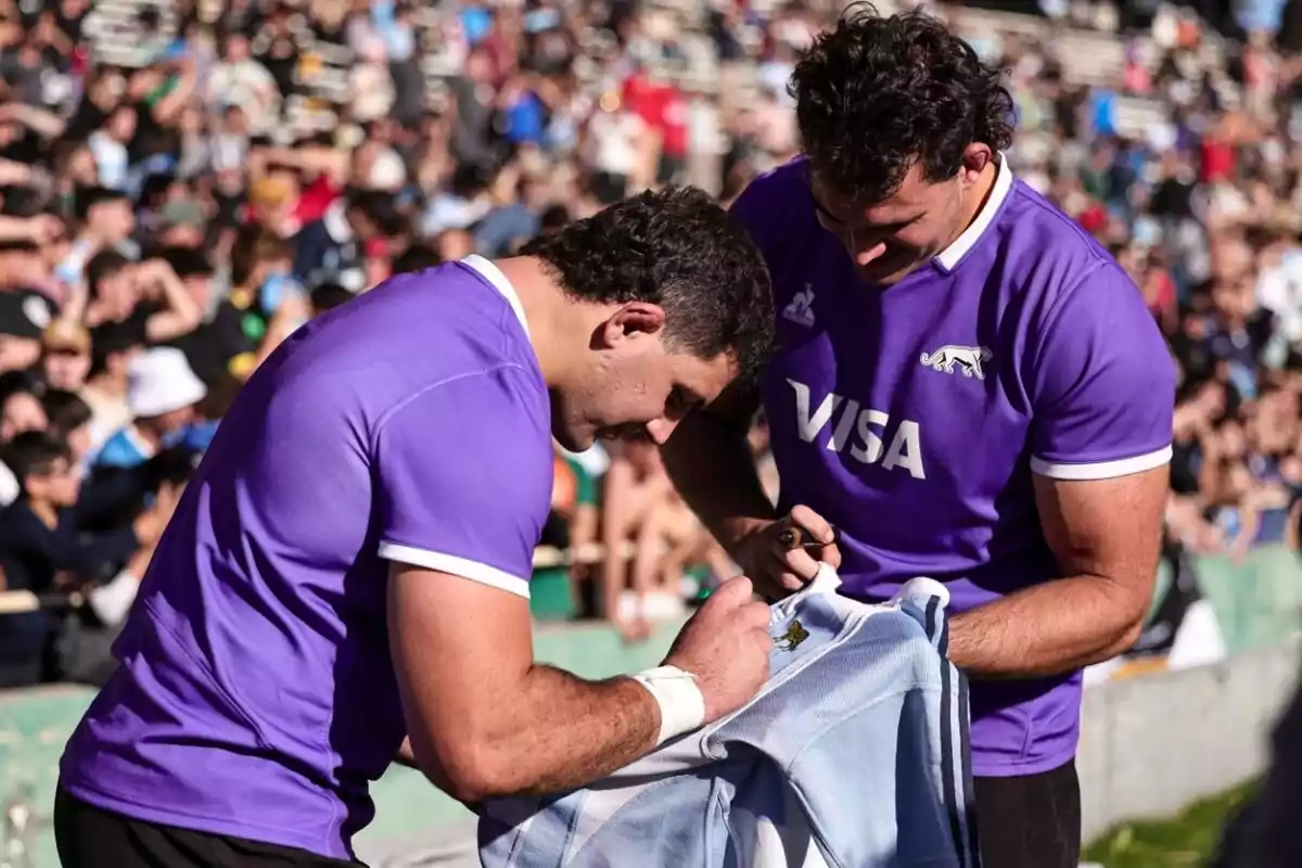 Two rugby players in purple jerseys sign a light blue and white jersey in a packed stadium.