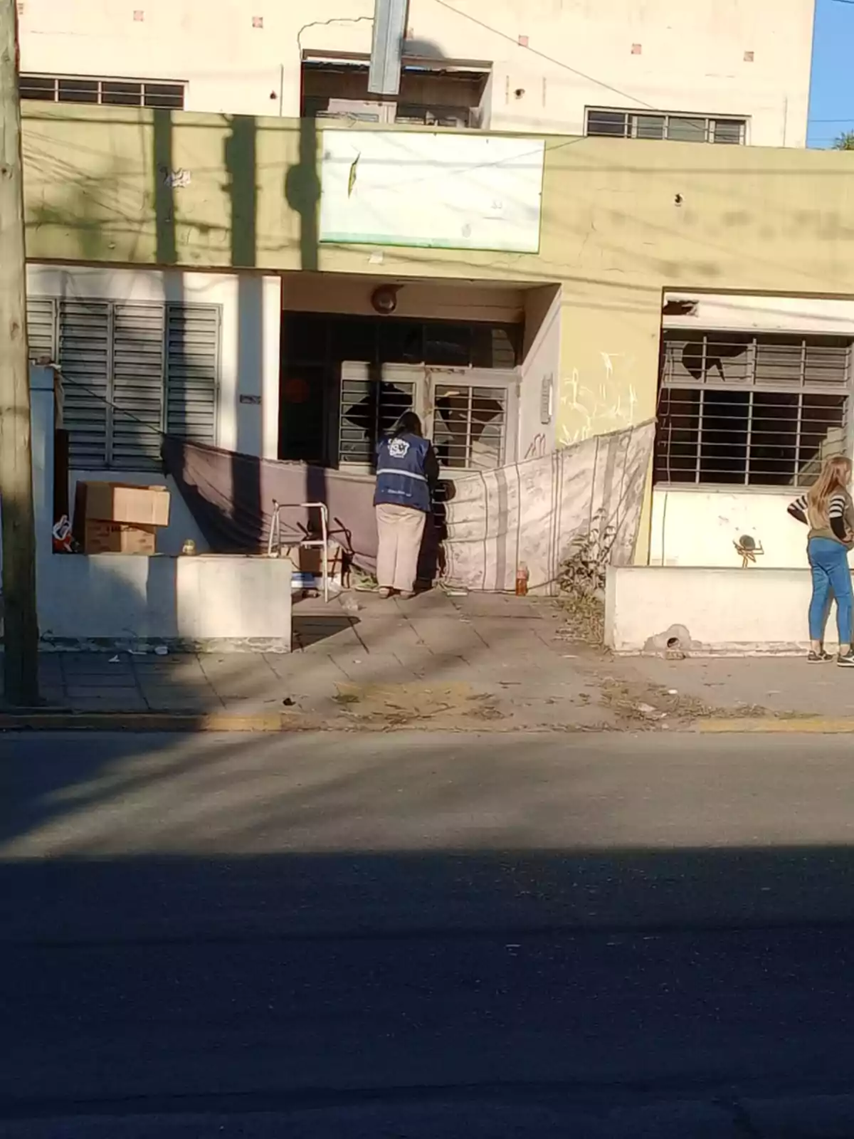 Persona con chaleco azul trabajando frente a un edificio con ventanas y cortinas improvisadas mientras otra persona está de pie cerca de la entrada Persona con chaleco azul trabajando frente a un edificio con ventanas y cortinas improvisadas mientras otra persona está de pie cerca de la entrada