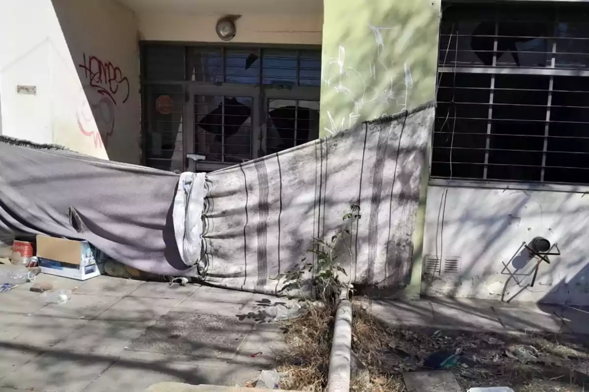 Fachada de un edificio abandonado con ventanas rotas, grafitis en la pared y mantas colgadas improvisando una especie de refugio, rodeado de basura y objetos tirados en la acera. Fachada de un edificio abandonado con ventanas rotas, grafitis en la pared y mantas colgadas improvisando una especie de refugio, rodeado de basura y objetos tirados en la acera.