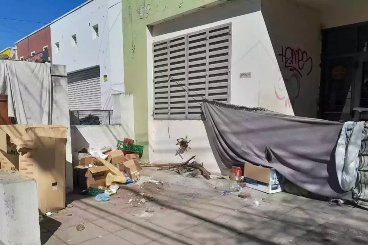 Una calle con basura, cajas de cartón, botellas y una manta colgada improvisando un refugio frente a un edificio con paredes blancas y verdes