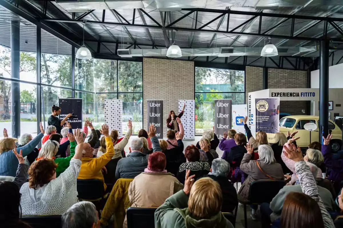 Un grupo de personas mayores participa en una charla interactiva en un salón moderno con ventanales, muchas de ellas levantan la mano mientras una mujer al frente dirige la actividad y se observan banners de Uber y un auto antiguo amarillo al fondo. Un grupo de personas mayores participa en una charla interactiva en un salón moderno con ventanales, muchas de ellas levantan la mano mientras una mujer al frente dirige la actividad y se observan banners de Uber y un auto antiguo amarillo al fondo.