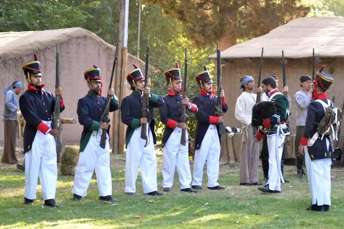 Grupo de hombres vestidos con uniformes militares históricos sosteniendo rifles en formación sobre césped con casas de adobe al fondo