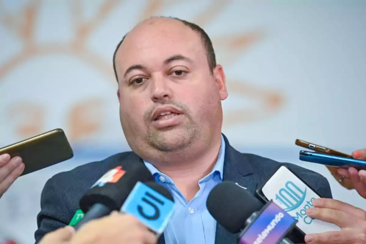 A bald man in a light blue jacket is being interviewed by several media outlets holding microphones and phones in front of him.