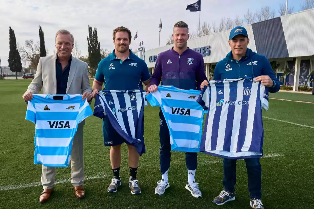 Four men pose on a grass field holding rugby and soccer jerseys in sky blue and white and blue and white colors.