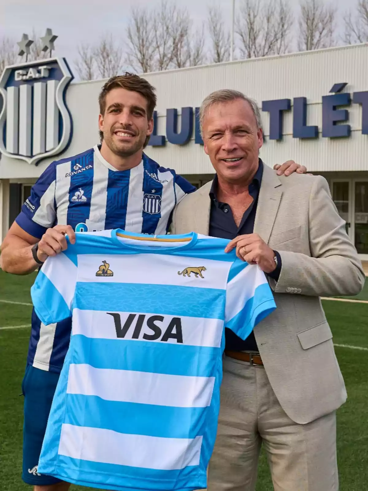 Two men smile and pose on a soccer field holding a light blue and white rugby jersey in front of a building with the crest and name of a sports club. Two men smile and pose on a soccer field holding a light blue and white rugby jersey in front of a building with the crest and name of a sports club.