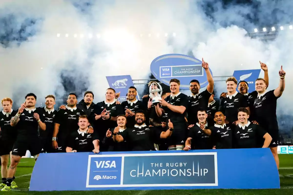 Jugadores de rugby celebran con un trofeo tras ganar el torneo The Rugby Championship, posando juntos frente a un cartel del campeonato con humo de fondo. Jugadores de rugby celebran con un trofeo tras ganar el torneo The Rugby Championship, posando juntos frente a un cartel del campeonato con humo de fondo.
