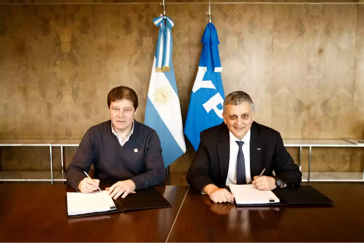 Dos hombres sentados en una mesa firmando documentos con una bandera de Argentina y una bandera de YPF al fondo Dos hombres sentados en una mesa firmando documentos con una bandera de Argentina y una bandera de YPF al fondo
