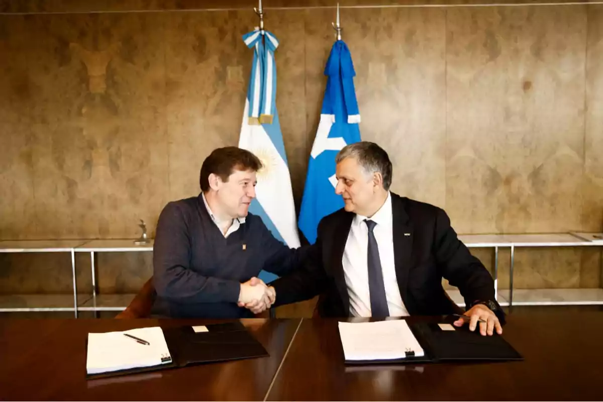 Dos hombres se dan la mano sentados en una mesa con carpetas y documentos, al fondo hay dos banderas, una de Argentina y otra azul