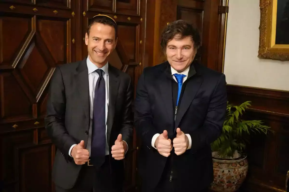 Dos hombres vestidos de traje sonríen y muestran ambos pulgares hacia arriba en una habitación elegante con paredes de madera y una planta decorativa al fondo Dos hombres vestidos de traje sonríen y muestran ambos pulgares hacia arriba en una habitación elegante con paredes de madera y una planta decorativa al fondo