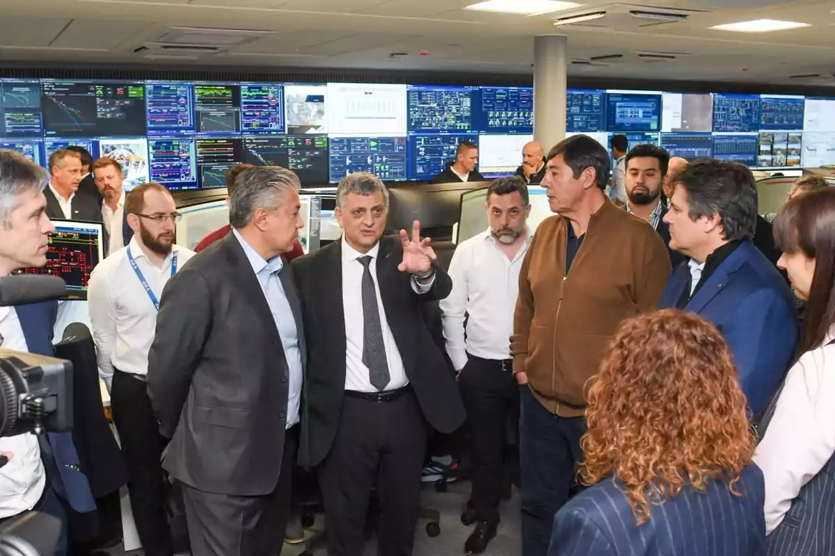 A group of people dressed formally are talking in a control room with multiple screens filled with charts and data in the background.