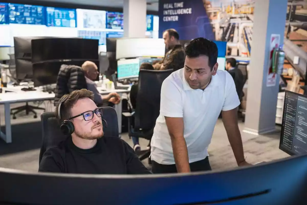 Two men working together in a modern office with several screens and monitors displaying data in the background Two men working together in a modern office with several screens and monitors displaying data in the background