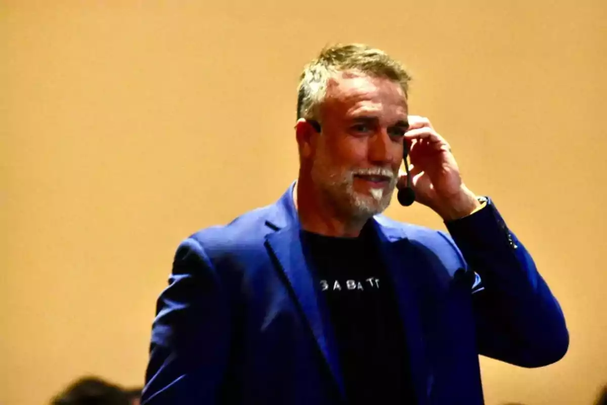 Short-haired, gray-bearded man wearing a headset microphone and a blue blazer while smiling