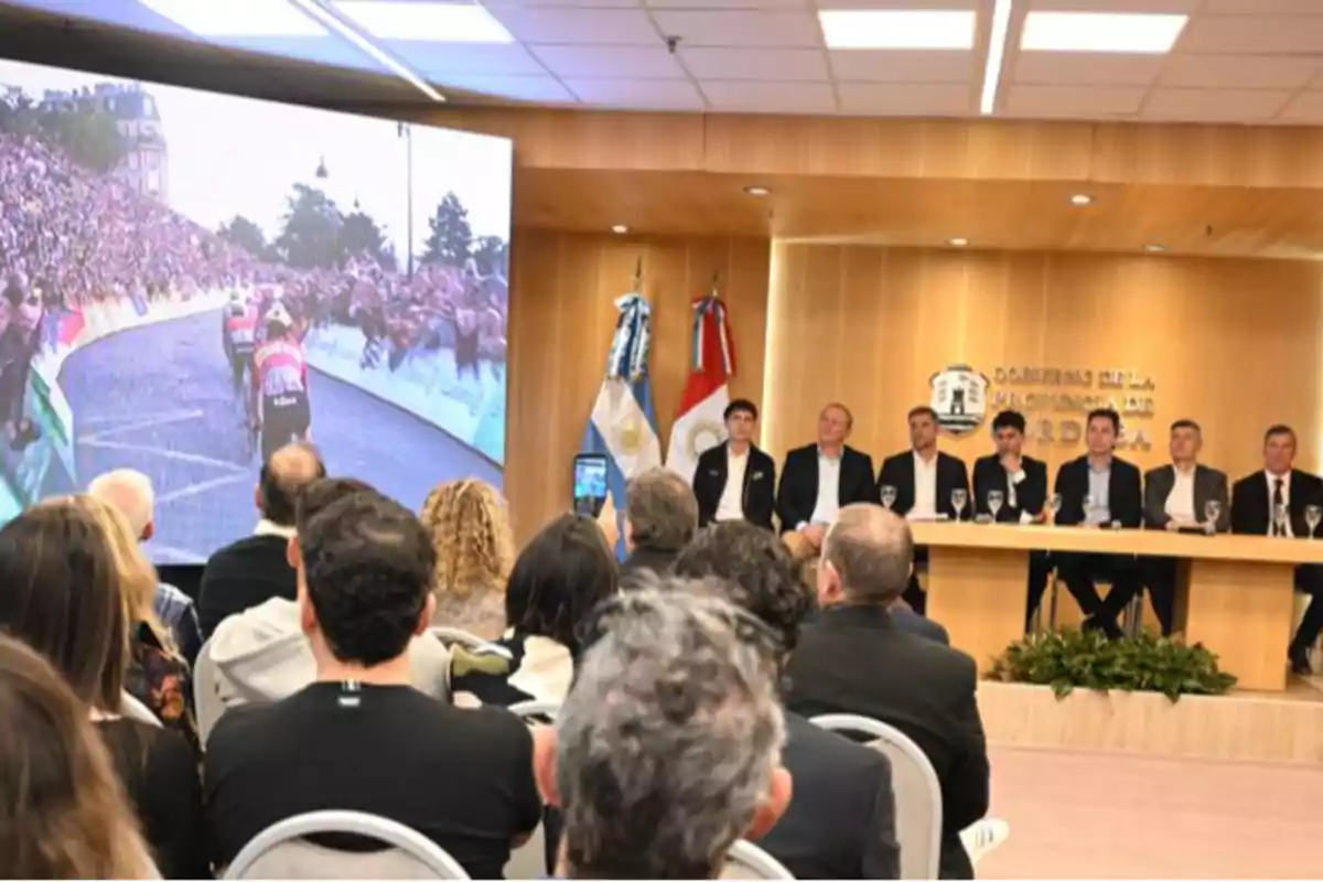 People seated in an auditorium watch a large screen showing a cycling race while a group of men sit at a main table in front of official flags. People seated in an auditorium watch a large screen showing a cycling race while a group of men sit at a main table in front of official flags.