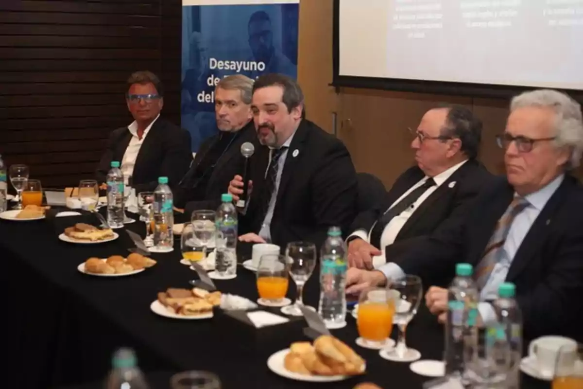 Five men in suits are seated at a table during a working breakfast, with a microphone, water, juice, and food on the table, while in the background a presentation screen and a blue sign can be seen.