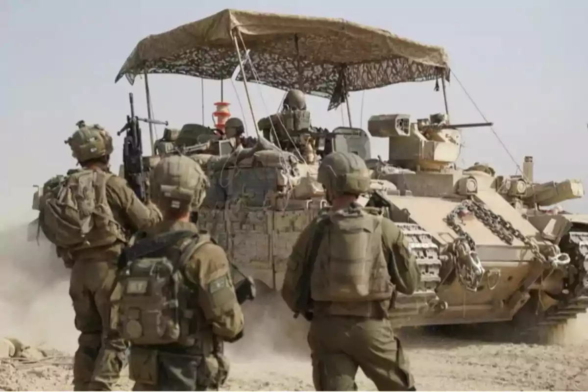 Soldados con equipo militar caminan junto a un vehículo blindado en un terreno arenoso bajo el sol Soldados con equipo militar caminan junto a un vehículo blindado en un terreno arenoso bajo el sol