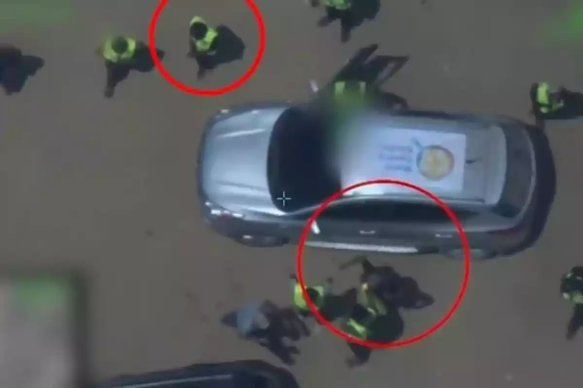 Vista aérea de un automóvil gris rodeado por varias personas con chalecos reflectantes, dos de las cuales están dentro de círculos rojos