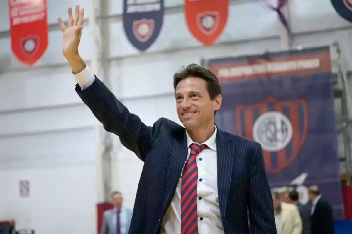 Man in a suit and tie smiles and waves, raising his hand in a gym decorated with sports pennants.