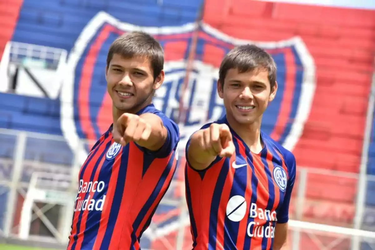Two players in San Lorenzo uniforms smile and point toward the camera in a stadium with the club’s colors and crest in the background. Two players in San Lorenzo uniforms smile and point toward the camera in a stadium with the club’s colors and crest in the background.