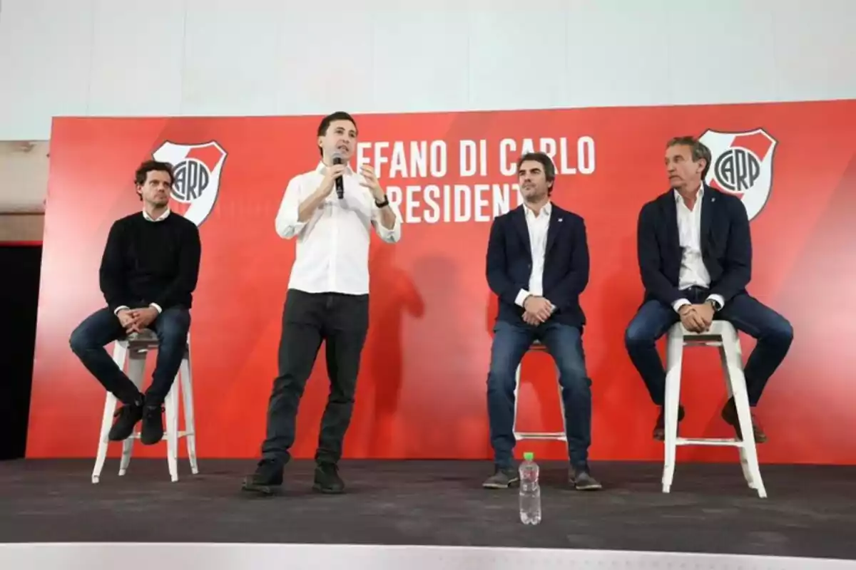 Four men on a stage with a red background and the River Plate crest, one of them standing and speaking into a microphone while the other three are seated on high stools. Four men on a stage with a red background and the River Plate crest, one of them standing and speaking into a microphone while the other three are seated on high stools.