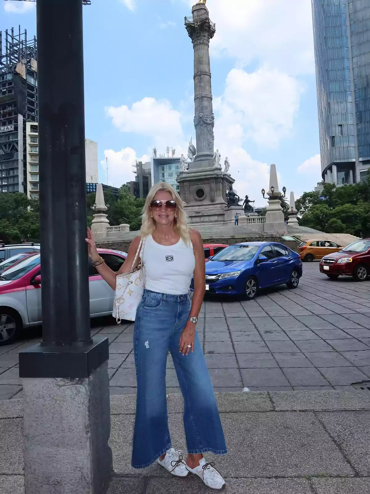 Mujer rubia con gafas de sol y ropa casual posando frente al Ángel de la Independencia en la Ciudad de México rodeada de autos y edificios modernos Mujer rubia con gafas de sol y ropa casual posando frente al Ángel de la Independencia en la Ciudad de México rodeada de autos y edificios modernos