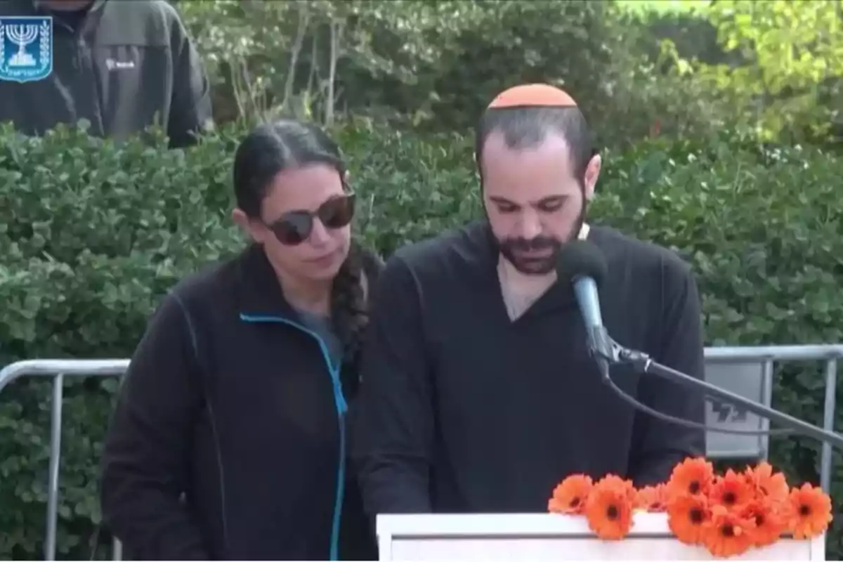 Two people standing in front of a microphone, one reading and the other showing support, with orange flowers decorating the lectern and green bushes in the background. Two people standing in front of a microphone, one reading and the other showing support, with orange flowers decorating the lectern and green bushes in the background.