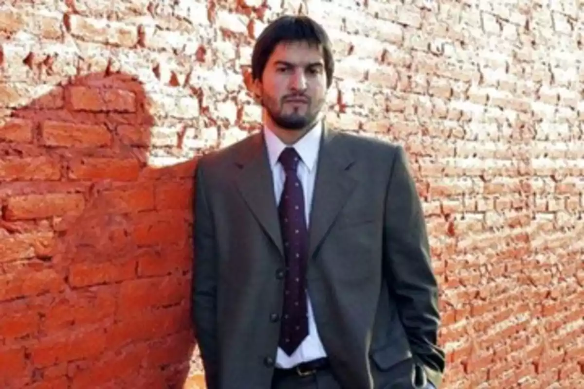 Man with a beard and short hair dressed in a formal suit leaning against a red brick wall