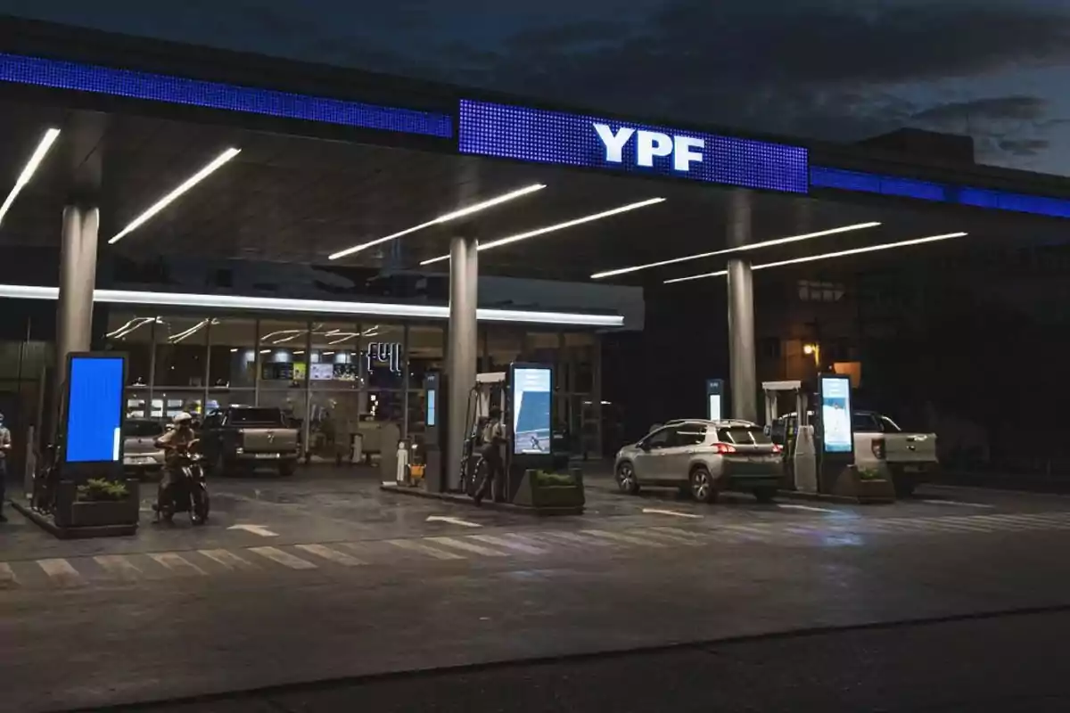 Estación de servicio YPF iluminada durante la noche con autos y una moto cargando combustible Estación de servicio YPF iluminada durante la noche con autos y una moto cargando combustible