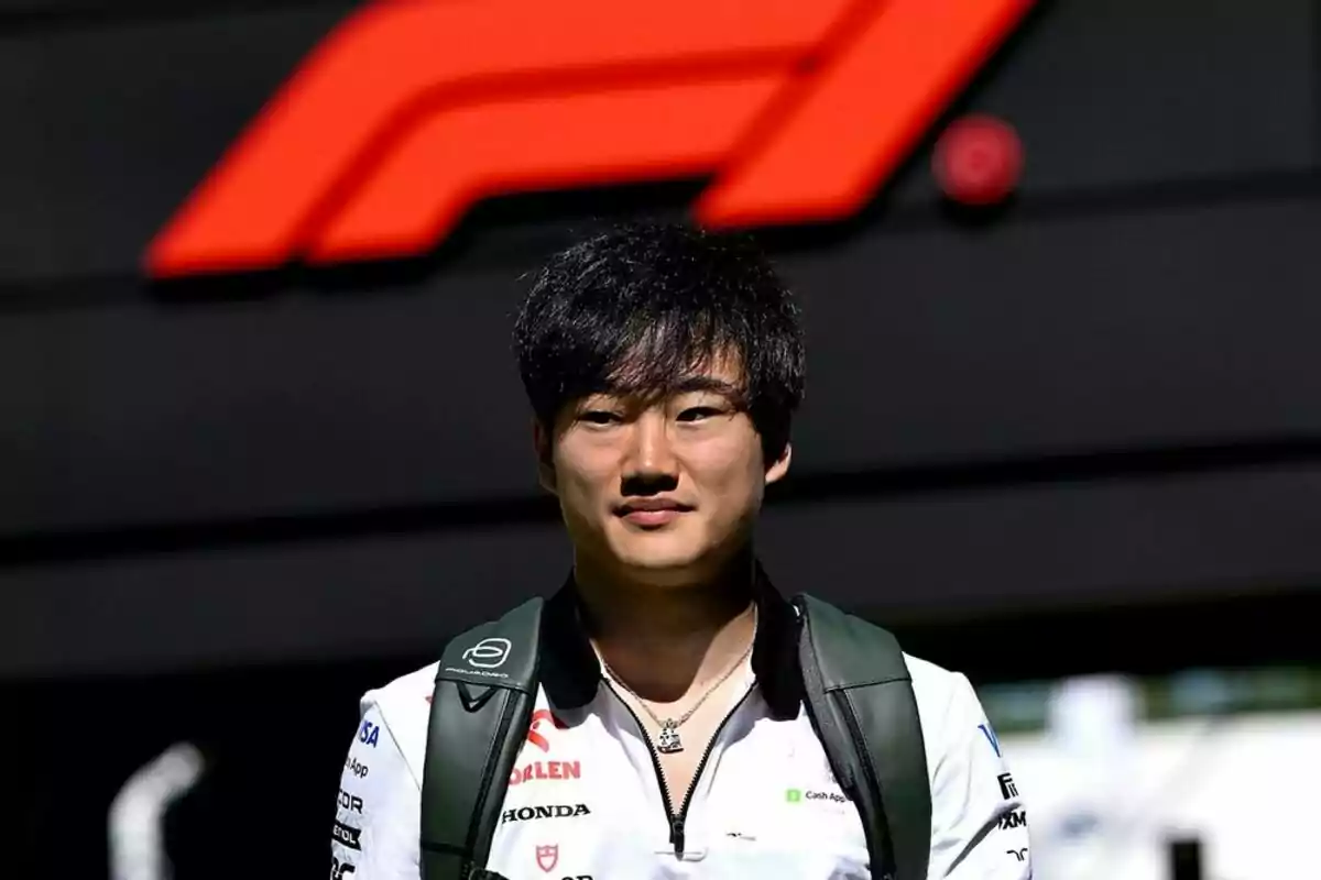 Joven con camiseta blanca y mochila verde de pie frente a un logo grande de Fórmula 1 Joven con camiseta blanca y mochila verde de pie frente a un logo grande de Fórmula 1