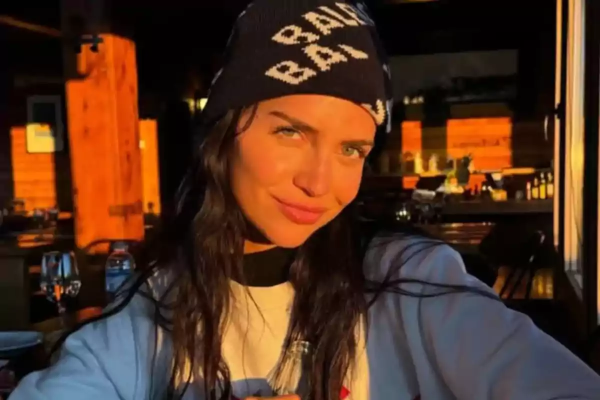 Mujer de cabello oscuro con gorro negro y suéter claro sentada en un restaurante iluminado por la luz del atardecer