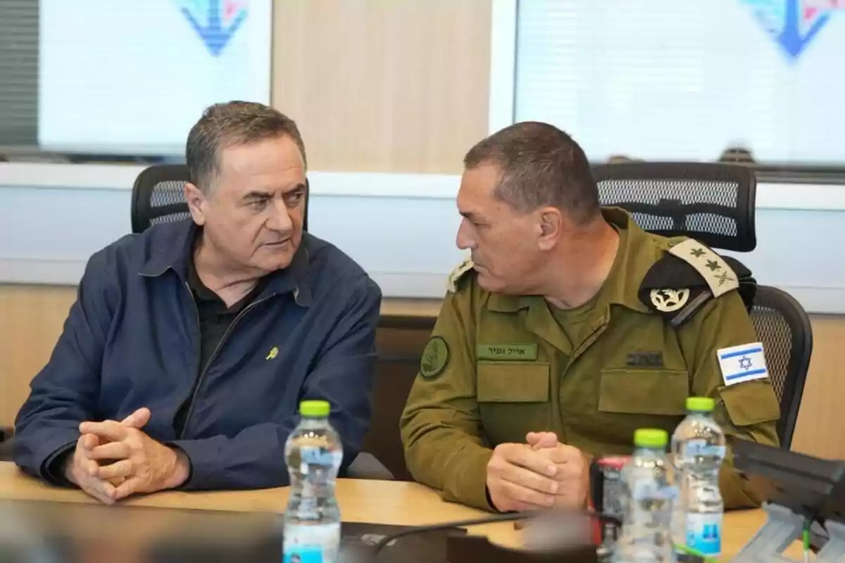 Dos hombres sentados en una mesa conversan seriamente, uno de ellos lleva uniforme militar israelí y el otro viste ropa civil azul, sobre la mesa hay botellas de agua y latas