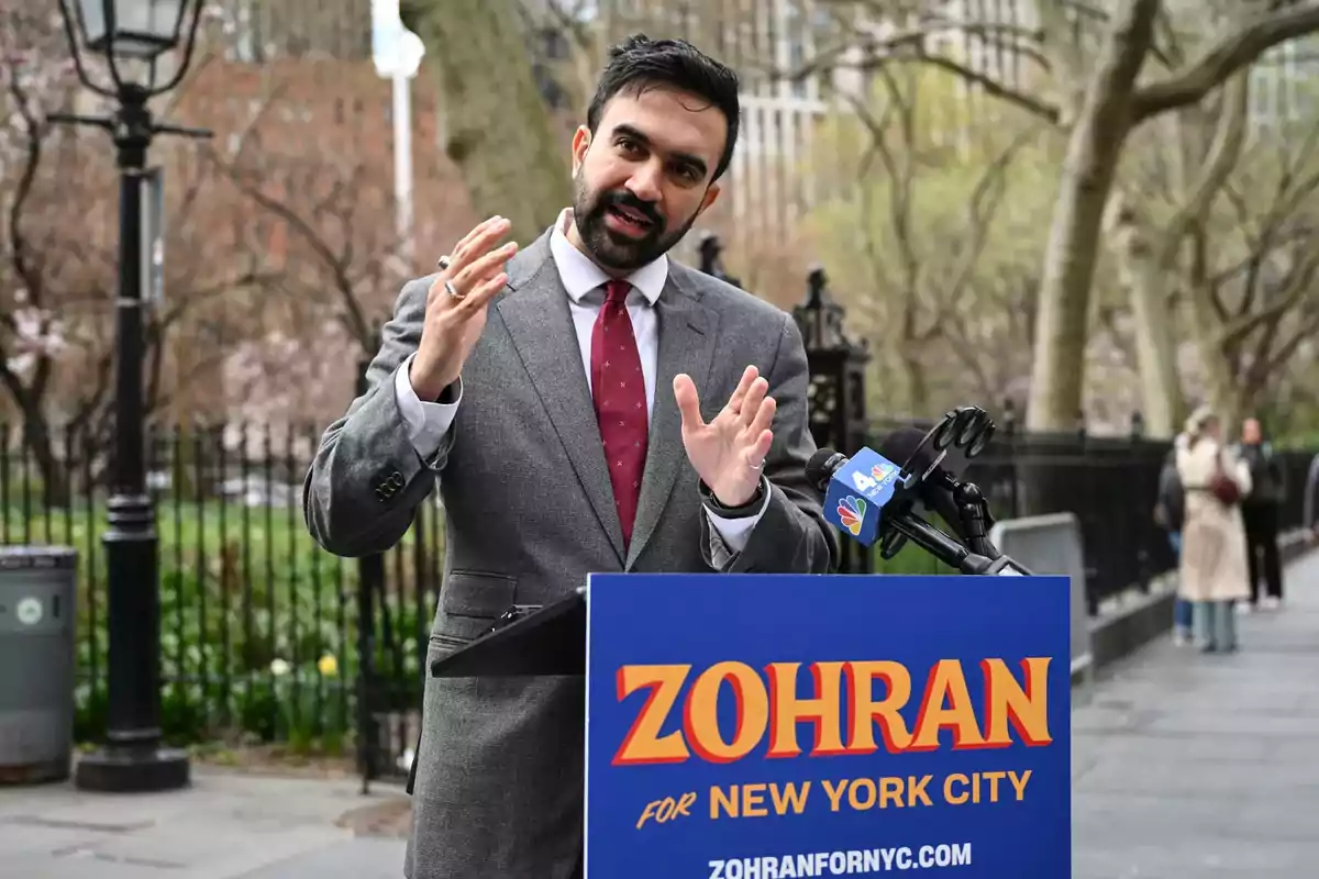Hombre con barba y traje gris hablando en un podio azul con el nombre Zohran en letras grandes durante un evento al aire libre en un parque