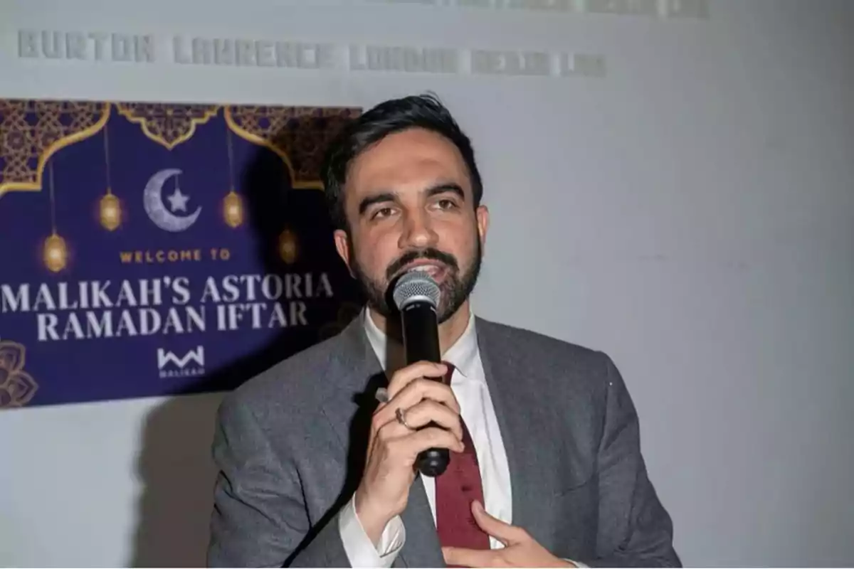 Hombre con barba y traje gris hablando por micrófono durante un evento de Ramadan Iftar con un cartel de fondo que dice Malikah's Astoria Ramadan Iftar