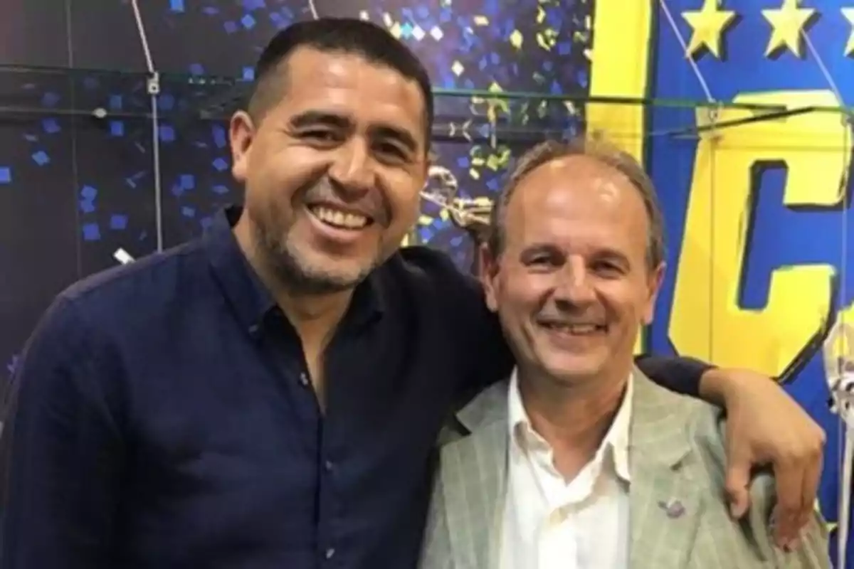 Two men smile, hugging in front of a blue and yellow background with stars and large letters. Two men smile, hugging in front of a blue and yellow background with stars and large letters.