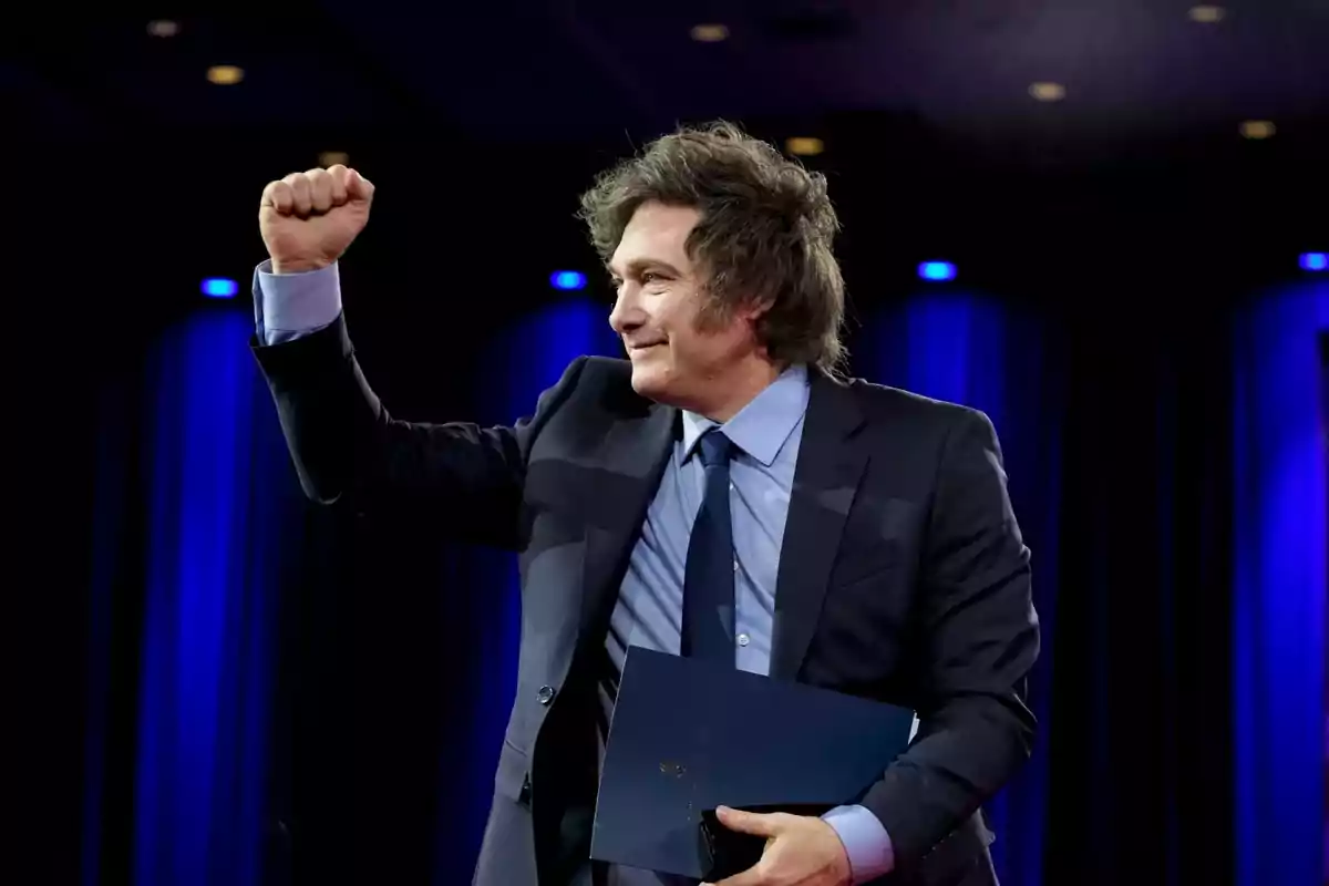 Disheveled-haired man in a blue suit raising his right fist in triumph against a background with blue lights