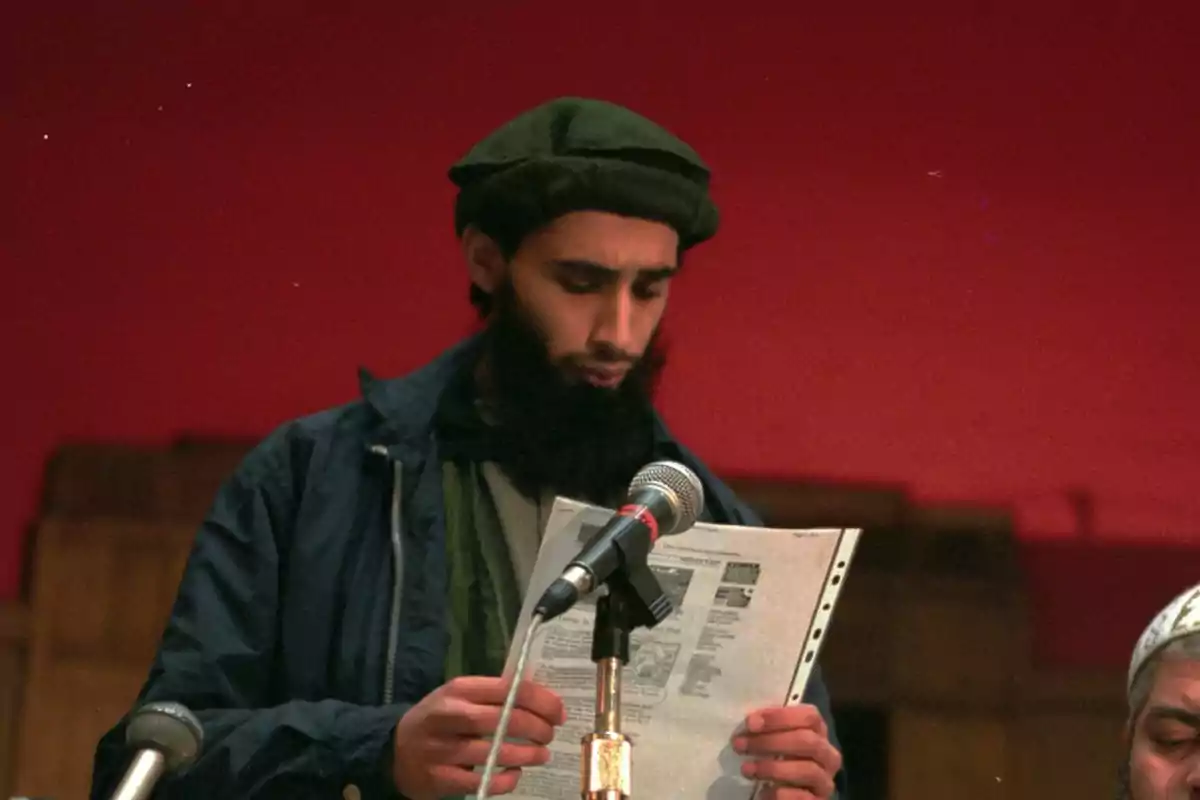 Hombre con barba y gorra leyendo un documento frente a un micrófono sobre un fondo rojo
