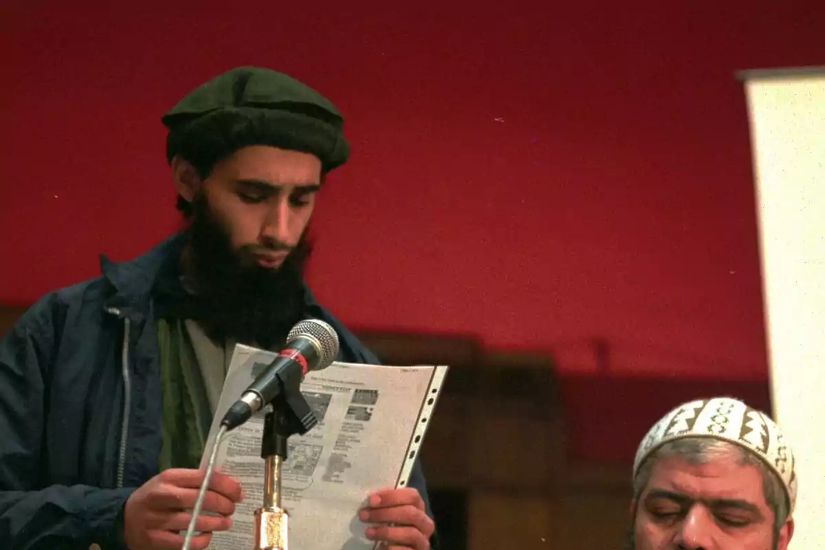 Hombre con barba y gorro verde leyendo un documento frente a un micrófono mientras otra persona está sentada a su lado Hombre con barba y gorro verde leyendo un documento frente a un micrófono mientras otra persona está sentada a su lado