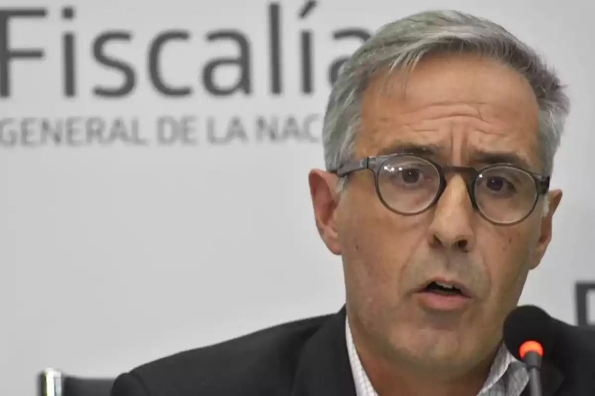 Hombre de cabello canoso y gafas hablando frente a un micrófono con un fondo que tiene el texto Fiscalía General de la Nación.