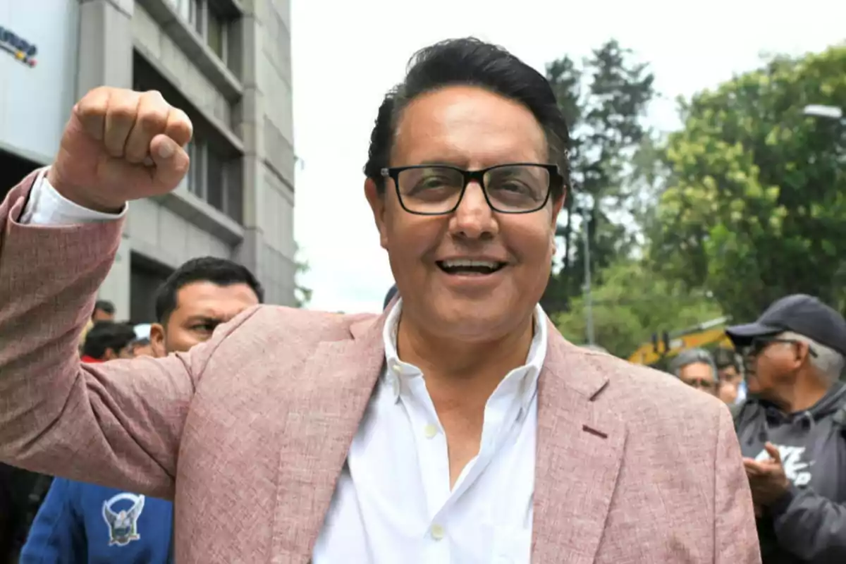 Man with glasses and a pink blazer smiles and raises his fist in celebration while surrounded by people outdoors