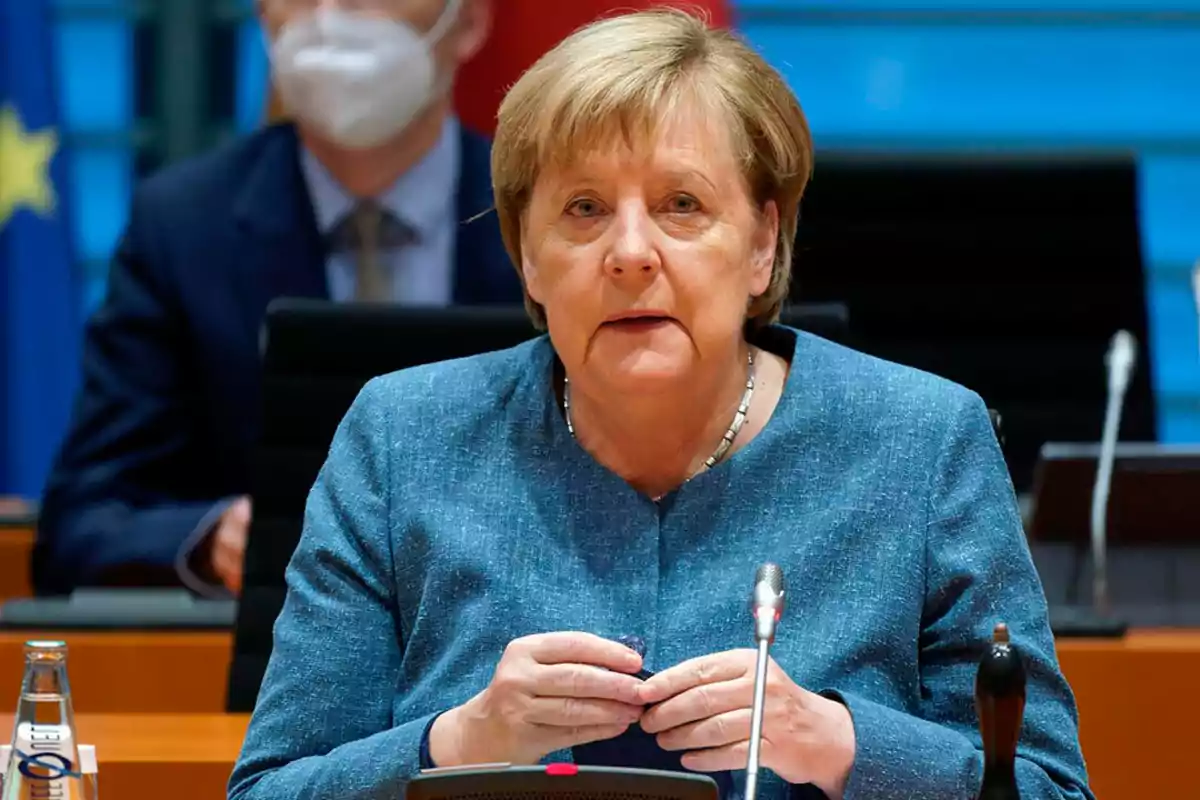 Blonde, short-haired woman sitting at a conference table with microphones and a water bottle, wearing a blue blazer, with a person wearing a face mask and blurred flags in the background.