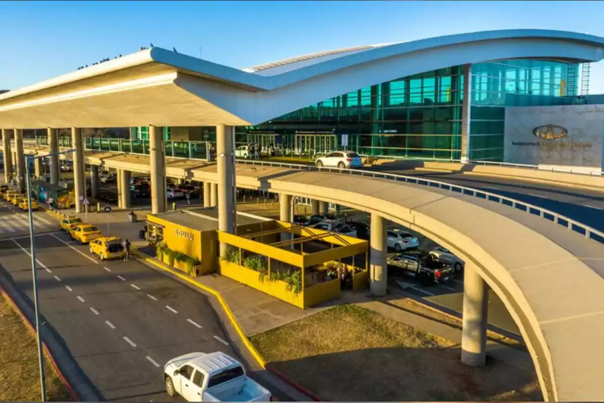 Exterior view of a modern airport with a curved roof structure, yellow taxis parked, and an elevated vehicle access area